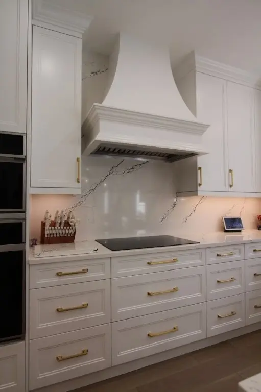 Modern kitchen stove area with white cabinets, gold handles, a white range hood, marble backsplash with gray veining, a black cooktop, and a knife block on the counter beside a small digital screen device.