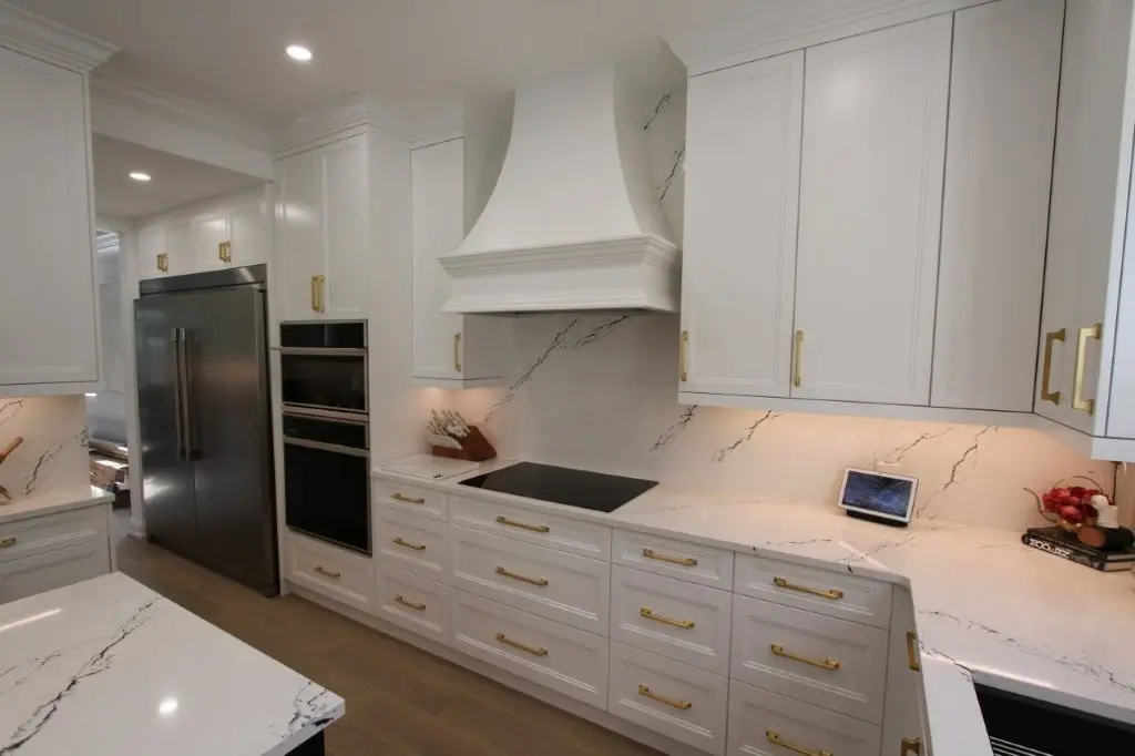 Modern kitchen with white cabinetry, gold handles, marble countertops and backsplash with black veining, stainless steel double-door refrigerator, built-in oven, electric cooktop under a white range hood, and wood flooring.