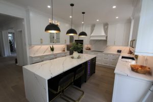 Modern kitchen with white marble countertops and backsplash, white cabinetry with gold handles, three black pendant lights over a large island with black seating and built-in wine cooler, light wood flooring, and various plants and kitchen accessories.