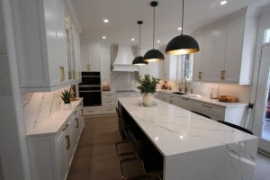 Modern white kitchen with marble countertops and backsplash, gold handles, a large island with black chairs, three pendant lights, and wooden flooring.