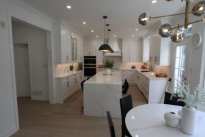 Modern kitchen with white cabinets, marble countertops, a large marble island with black chairs, pendant lighting, and a round white dining table with black chairs and white vases, brightened by natural light from a glass door.