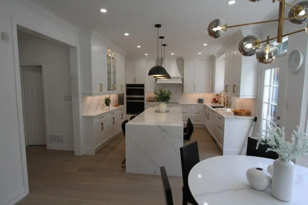 Modern kitchen with white cabinets, marble countertops, a large marble island with black chairs, pendant lighting, and a round white dining table with black chairs and white vases, brightened by natural light from a glass door.