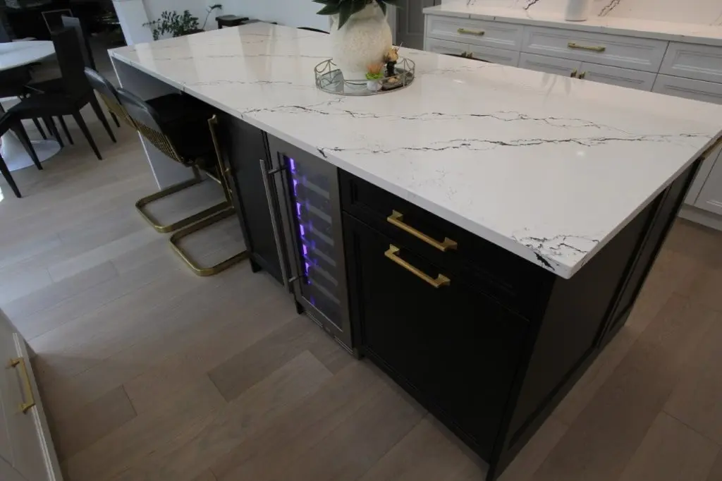 Modern kitchen island with black cabinetry, gold handles, and a white marble countertop with gray veining, featuring a built-in wine cooler with purple lighting and two black and gold bar stools on light wood flooring.