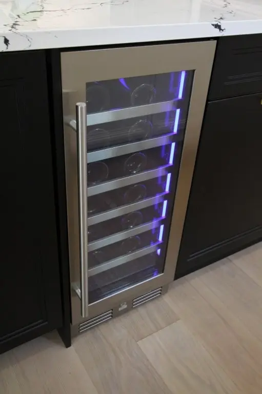 Built-in wine cooler with glass door and stainless steel frame, interior lit with blue light, showing multiple wine bottles on horizontal shelves inside a kitchen cabinet with black panels and white marble countertop