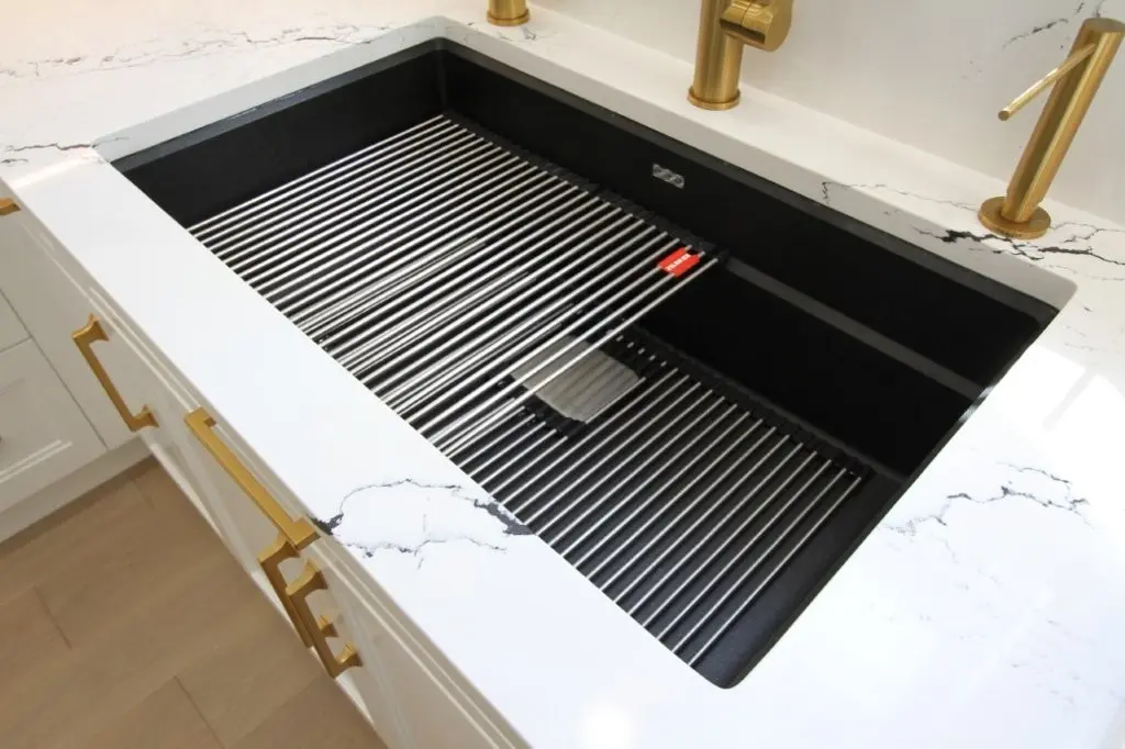 Modern black kitchen sink with a metal drying rack inside, set in a white countertop with subtle black veining and gold faucets and cabinet handles.