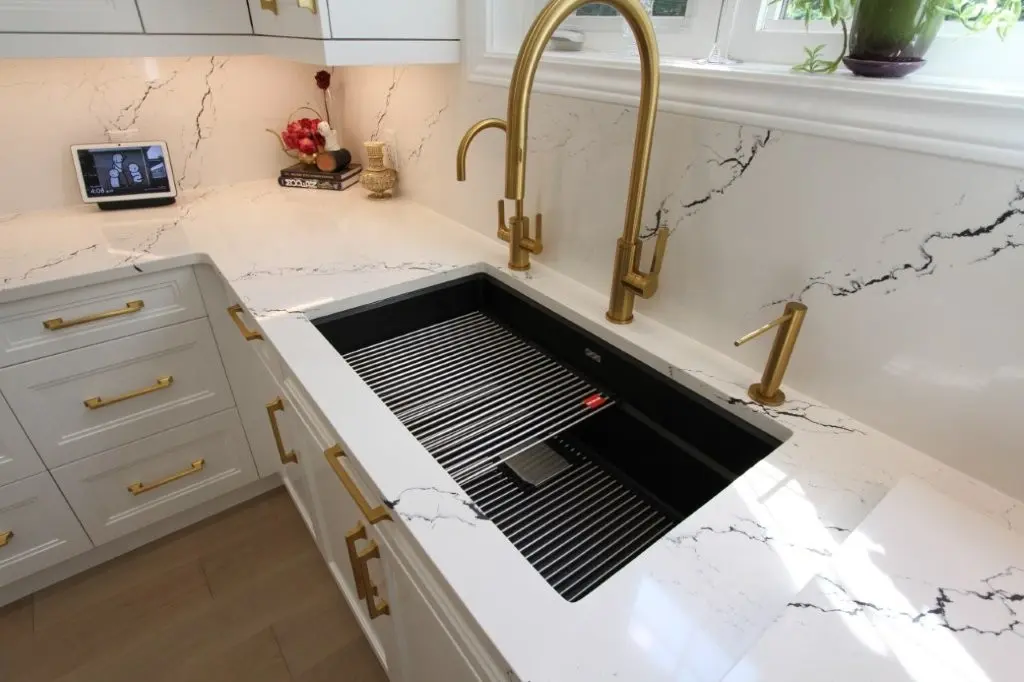 Modern kitchen sink area with a black undermount sink featuring a metal dish rack, marble-look white countertop and backsplash with black veining, gold faucet fixtures, white cabinetry with gold handles, and a smart display device on the corner of the counter.