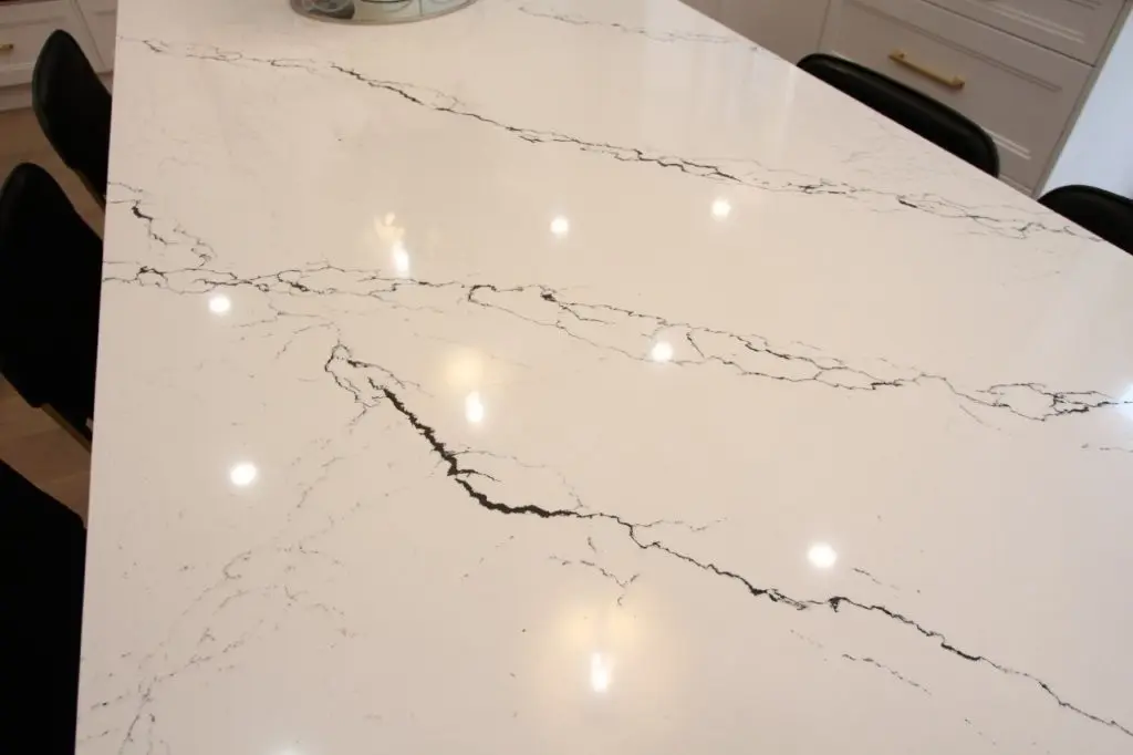 White marble tabletop with black veining and reflections of ceiling lights, surrounded by black chairs in a modern kitchen setting.