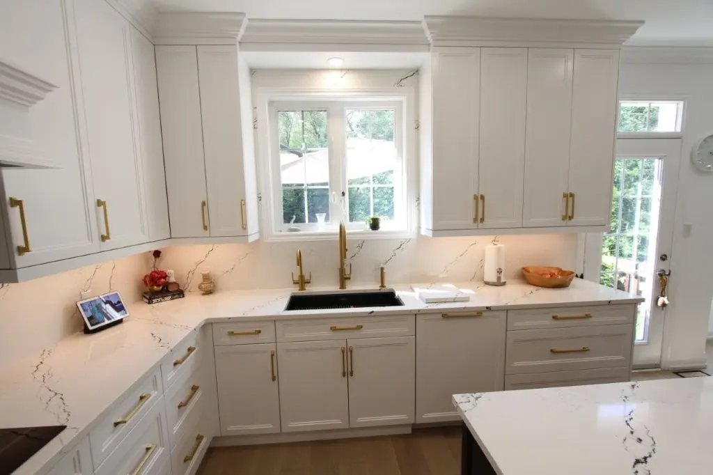Bright modern kitchen with white cabinetry, brass handles, white marble countertops with gray veining, a large window above a black sink with gold faucet, and light wood flooring.