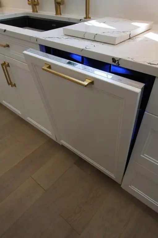 Modern kitchen dishwasher built into white cabinets with gold handles, partially open beneath a white marble countertop with black veining, next to a black sink and gold faucet fixtures, on a light wood floor.