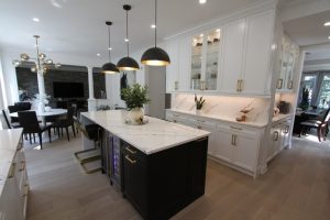 Modern kitchen with a white marble island countertop, black base cabinets with gold handles, three black pendant lights, and white wall cabinets with glass doors and under-cabinet lighting, adjacent to a dining area with a round table and black chairs.