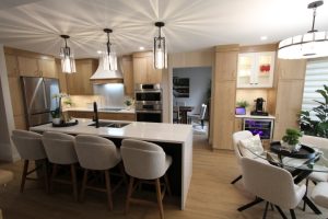Modern kitchen with light wooden cabinets, quartz island with black faucet and four upholstered bar stools, stainless steel appliances, pendant lights, and adjacent dining area with round glass table and beige chairs.
