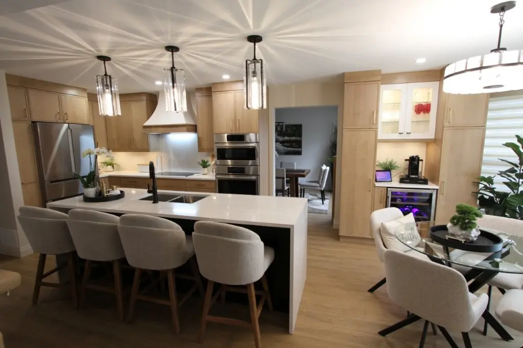 Modern kitchen with light wooden cabinets, quartz island with black faucet and four upholstered bar stools, stainless steel appliances, pendant lights, and adjacent dining area with round glass table and beige chairs.