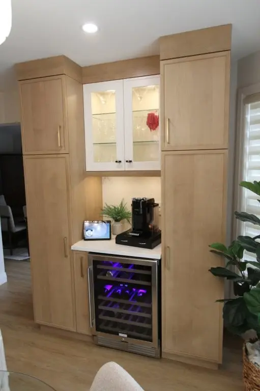 Wooden kitchen cabinetry with upper glass doors displaying glassware, a white countertop with a coffee maker, a tablet, and a small plant, and a built-in wine cooler with purple interior lighting beneath the counter, next to a window with blinds and a large green plant in a basket.