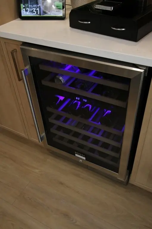 Built-in wine cooler with stainless steel frame and glass door, illuminated with purple interior light, installed under a light-colored countertop with wooden cabinets and light wood flooring.