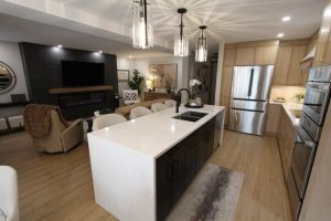 Modern open-concept kitchen and living room with a white quartz island, black cabinets, stainless steel appliances, light wooden cabinetry, and beige seating in the living area with a TV mounted above a fireplace.