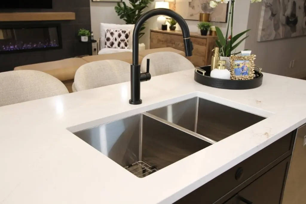 Modern kitchen island with a double stainless steel sink and a black high-arc faucet, featuring a white countertop and beige cushioned chairs, with decorative items and plants in the background.