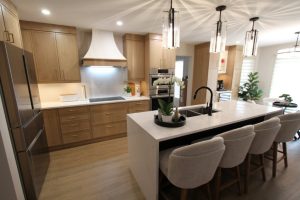 Modern kitchen with wooden cabinets, a white quartz countertop island with a black faucet, four upholstered bar stools, pendant lights above the island, a stainless steel refrigerator, built-in oven, and decorative plants on the island and near the window.