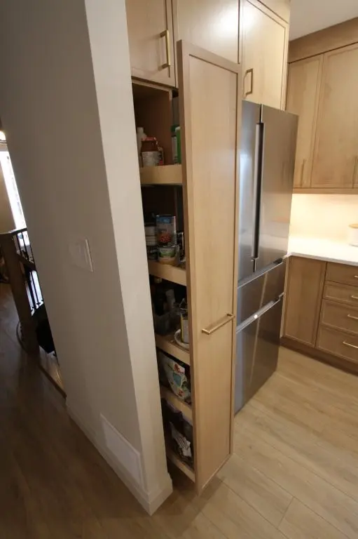 Tall kitchen pantry cabinet with pull-out shelves storing canned goods and jars next to a stainless steel refrigerator in a kitchen with light wood cabinetry and flooring.