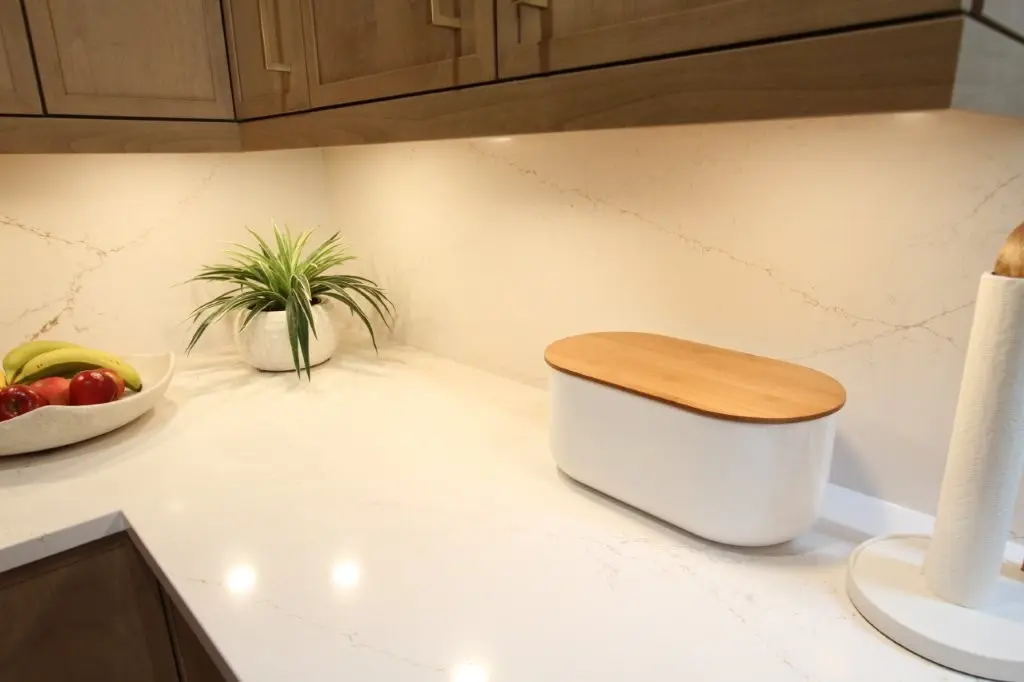 Clean kitchen countertop with a white container with a wooden lid, a paper towel holder, a small potted plant, and a bowl of fruit including bananas and apples, under wooden cabinets and bright lighting.