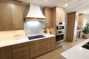 Modern kitchen with light wood cabinets, white countertops, built-in oven and microwave, induction cooktop with a white range hood, and a decorative plant on the counter.