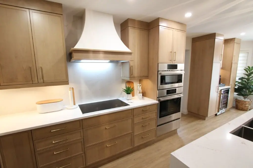Modern kitchen with light wood cabinets, white countertops, built-in oven and microwave, induction cooktop with a white range hood, and a decorative plant on the counter.