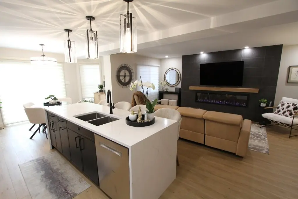 Modern open-concept living space with a kitchen island featuring a white quartz countertop and black cabinetry, beige leather sofa facing a wall-mounted TV above a linear electric fireplace, pendant lights with glass shades, and light wood flooring throughout.
