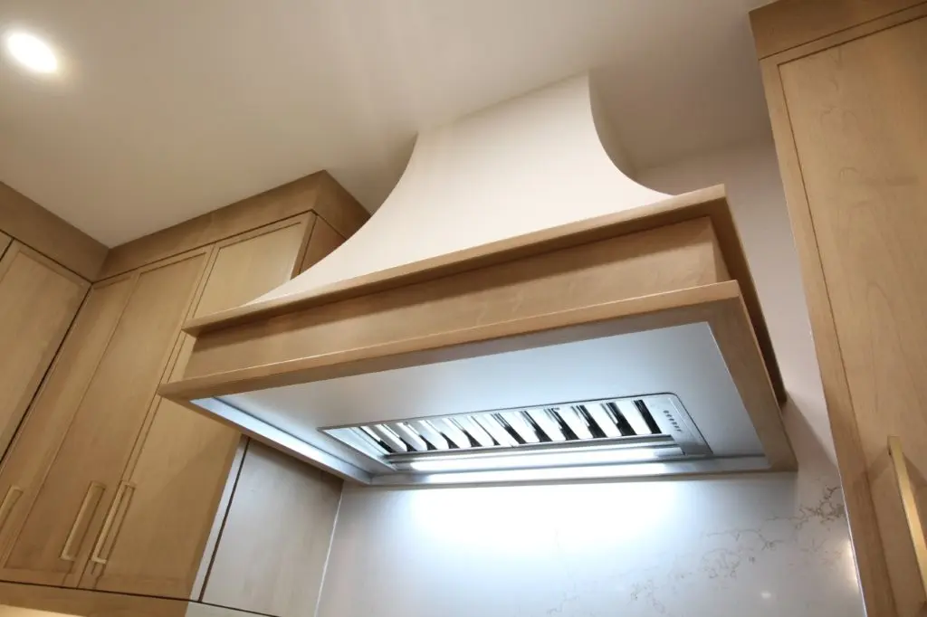 Underside view of a modern kitchen range hood with wooden trim and built-in lights, surrounded by wooden cabinets and a light-colored marble backsplash