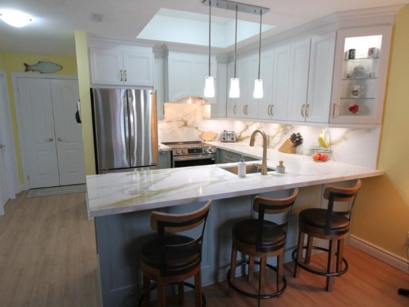 Bright kitchen with white and light blue cabinetry, quartz countertops, stainless steel refrigerator and oven, three wooden bar stools with black seats under a marble peninsula, and three pendant lights hanging above the countertop.