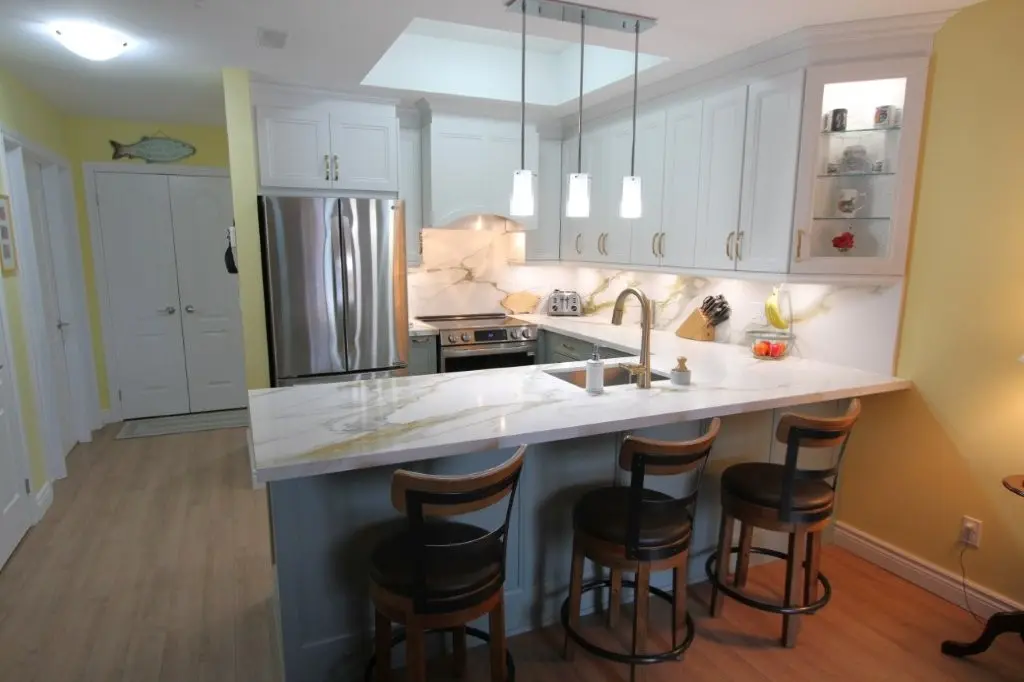 Bright kitchen with white and light blue cabinetry, quartz countertops, stainless steel refrigerator and oven, three wooden bar stools with black seats under a marble peninsula, and three pendant lights hanging above the countertop.