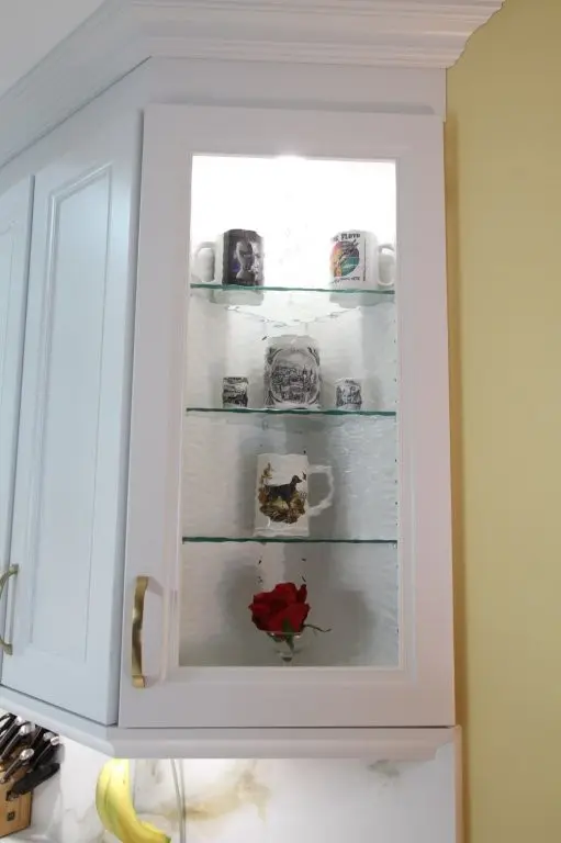 White kitchen cabinet with glass door containing three shelves displaying decorative mugs and a single red rose at the bottom shelf, with brass handles and a knife block visible below the cabinet.