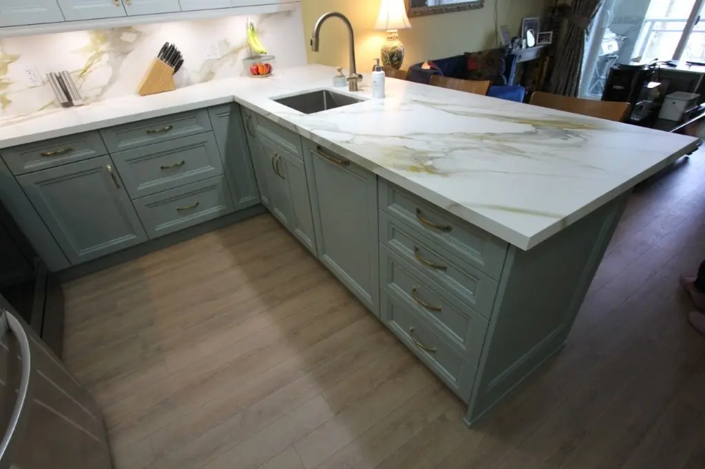 Modern kitchen with light green cabinetry and a white quartz countertop featuring gold and gray veins, a stainless steel sink with a brass faucet, a knife block, a bowl of fruit, and a decorated lamp in the background.