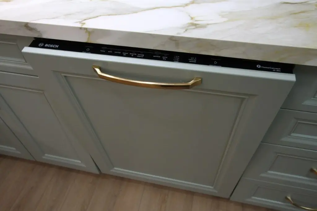 A Bosch built-in dishwasher with a gray panel and a gold handle, installed under a marble countertop in a kitchen with wood flooring and gray cabinetry.