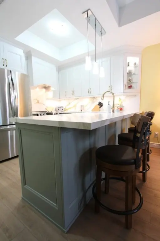Modern kitchen featuring a marble countertop island with three brown cushioned bar stools, white cabinetry, stainless steel refrigerator, and three pendant lights hanging from the ceiling.