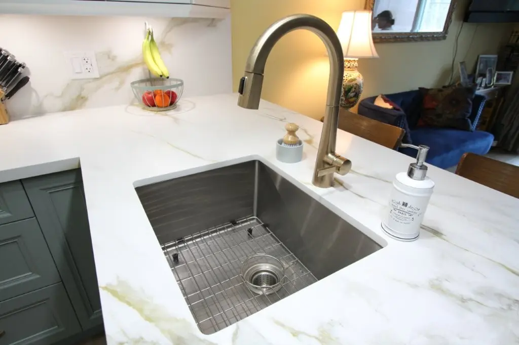 Modern kitchen sink with a stainless steel basin, gold-colored faucet, white quartz countertop, and a soap dispenser, with a fruit bowl and lamp visible in the background