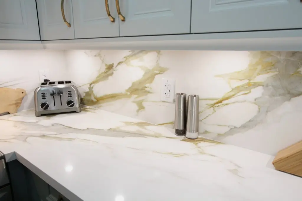 Modern kitchen countertop with white and gold veined quartz, featuring a stainless steel toaster, two metal salt and pepper grinders, and a light-colored cutting board, under white cabinets with gold handles and LED strip lighting.