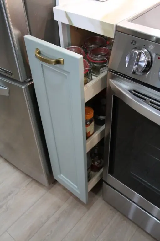 Narrow kitchen cabinet drawer pulled out between stainless steel refrigerator and oven, containing glass jars with red lids and spice containers.