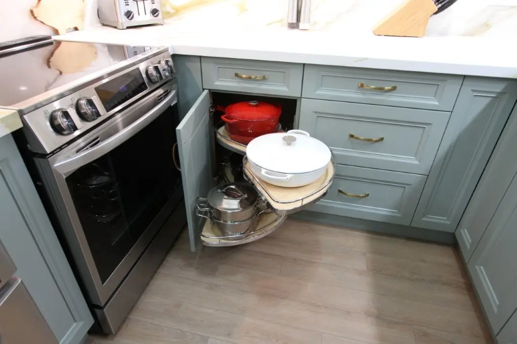Corner kitchen cabinet with a pull-out lazy Susan shelf holding pots, including a red pot and a white pot, next to a stainless steel oven and salt and pepper shakers on the countertop.