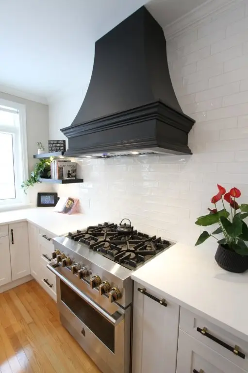 Modern white kitchen with stainless steel gas range, large black vent hood, white subway tile backsplash, and potted red anthuriums