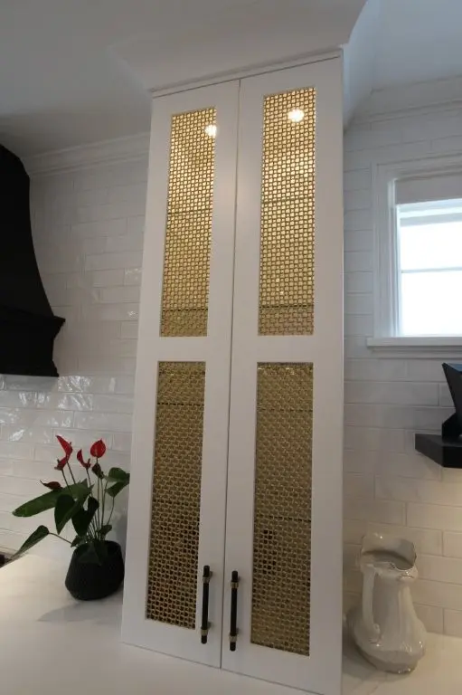 Tall white kitchen cabinet with brass honeycomb mesh inset panels, black handles, tile backsplash, plant and window nearby