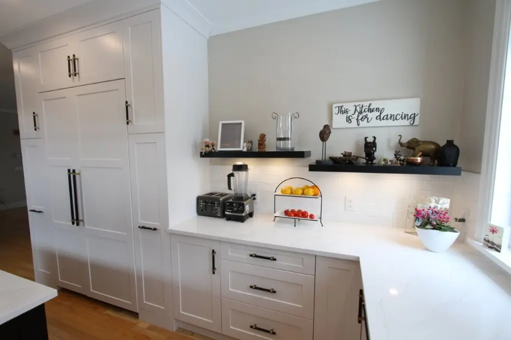 Bright modern kitchen corner with white shaker cabinets, panel ready fridge, quartz countertop, floating shelves with decor and a fruit stand by a window