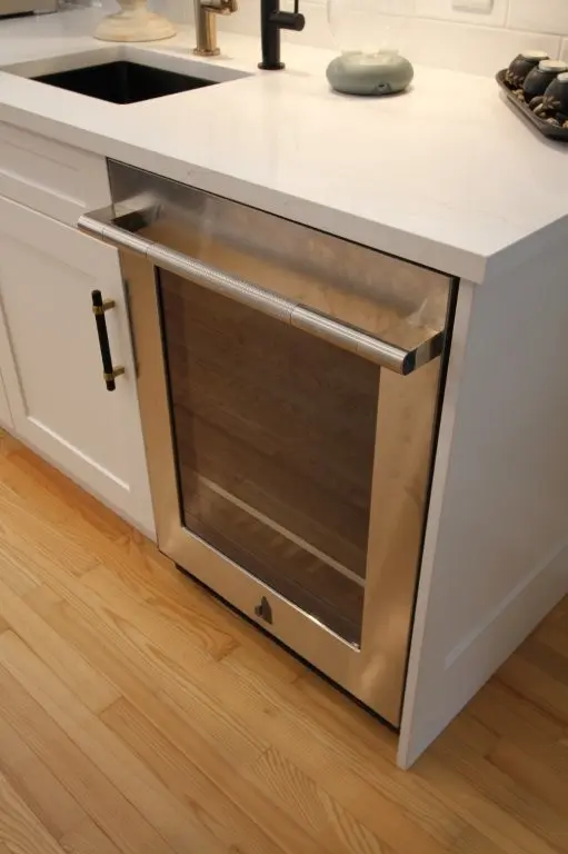 Built-in stainless steel wine fridge with glass door under a white countertop next to a black sink and white cabinetry.