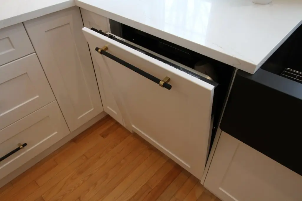 White kitchen cabinet panel ready dishwasher slightly ajar under a white quartz countertop, black handle with brass accents.