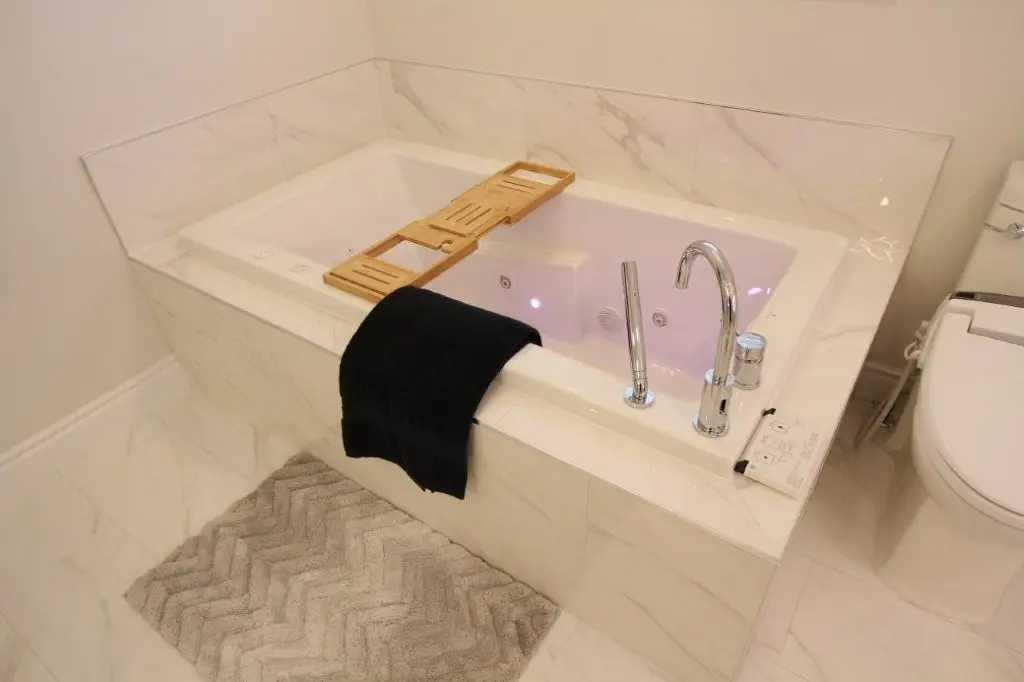 Modern white porcelain enclosure with a built in whirlpool tub, chrome faucet, wooden bath tray, black towel, and gray bath mat