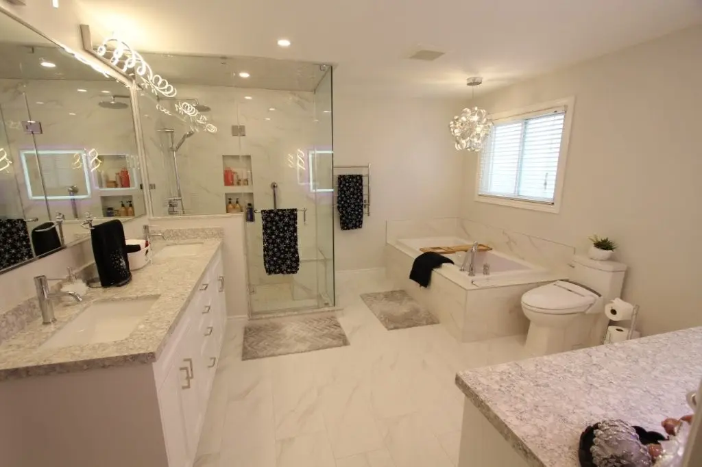 Bright modern bathroom with double sinks, glass shower, built in tub, and toilet with bidet seat in white marble-tone finishes.