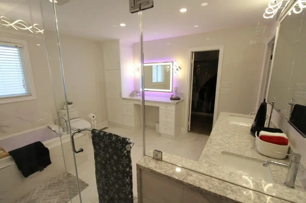 Spacious modern bathroom with glass shower, double-sink Quartz topped vanity, bathtub, illuminated mirror with purple backlight, and towels.