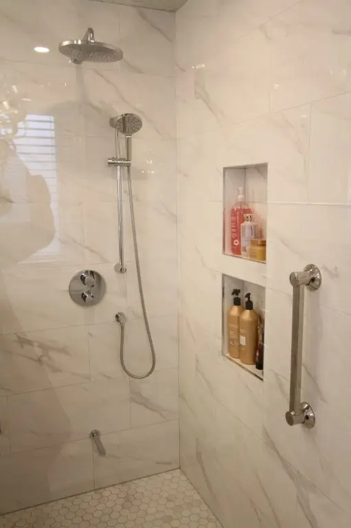 Shower with chrome rain showerhead and handheld wand, built-in recessed shelves holding bottles, marble-look porcelain tiled walls and hexagon floor tiles, chrome grab bar.