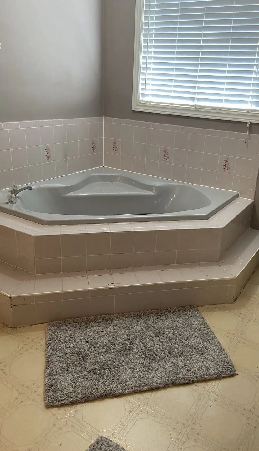 before picture of corner built-in bathtub with tiled surround and steps, pale gray walls, window with blinds above, and a gray bath mat on the floor