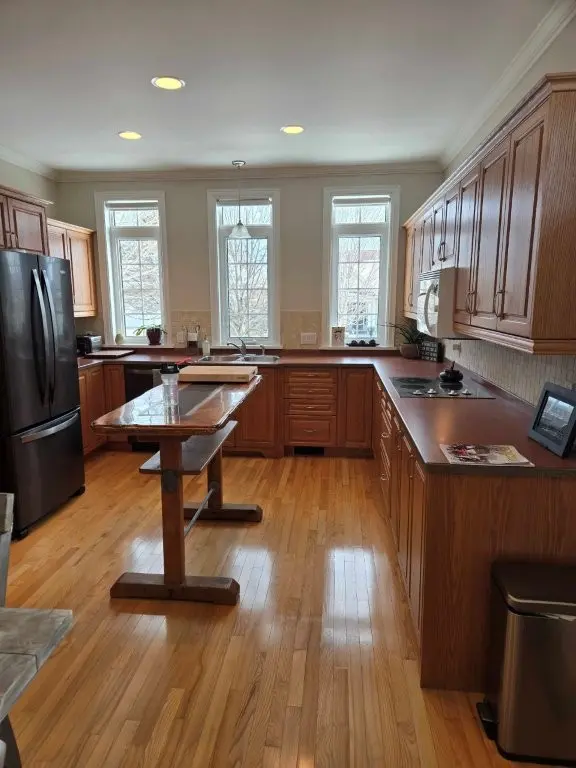 Before renovation picture. Kitchen with wooden cabinets, center island table, hardwood floors, stainless fridge and built-in stove.