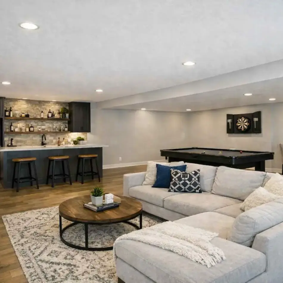 Spacious finished basement with a large light-gray sectional, round wood coffee table, area rug, wall-mounted TV, wet bar with three stools, and a pool table. Spacious finished basement with a large light-gray sectional, round wood coffee table, area rug, wall-mounted TV, wet bar with three stools, and a pool table.