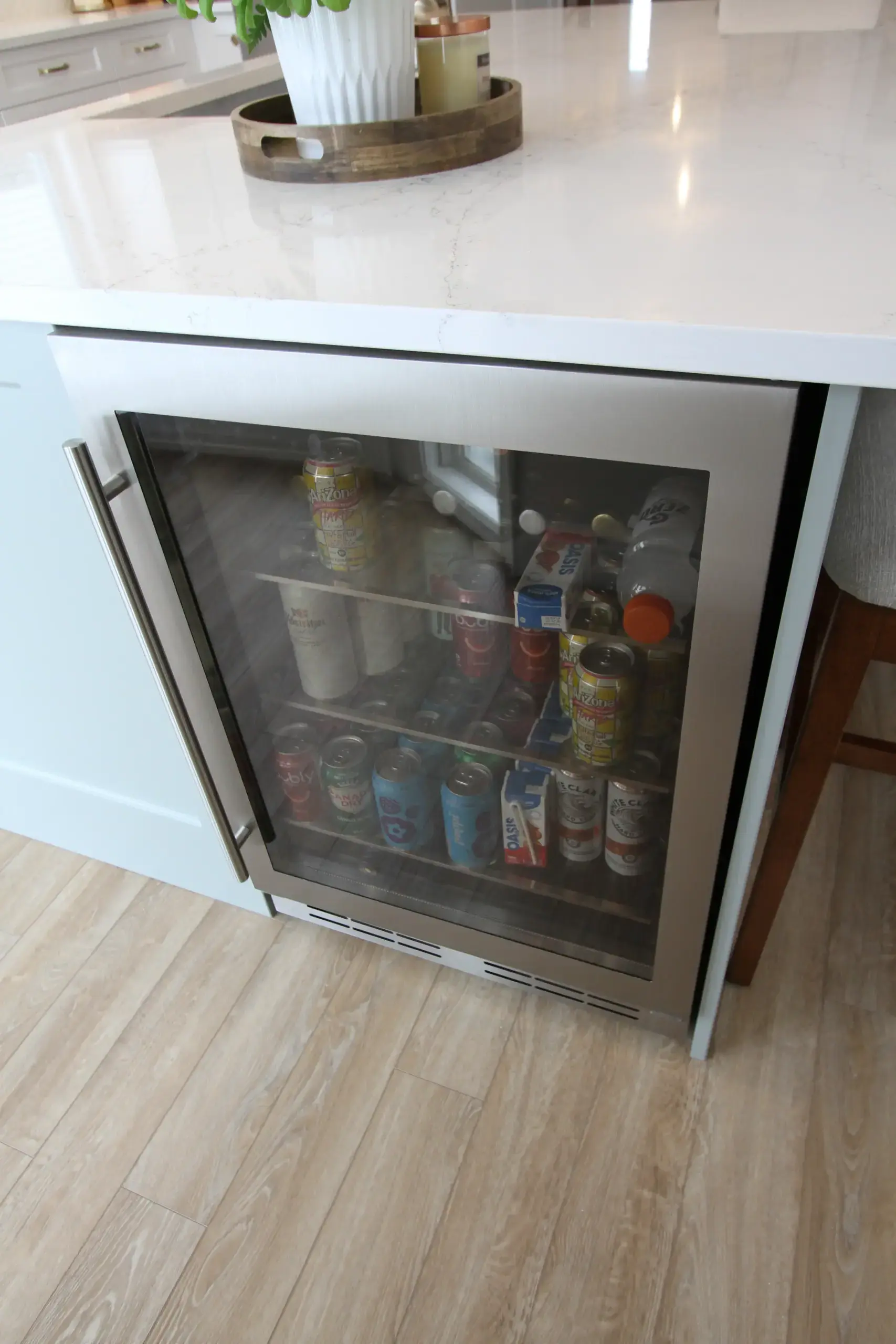 Kitchen island with a glass-door beverage fridge stocked with cans and bottles underneath a white countertop